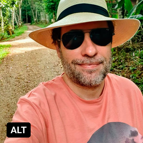 Selfie de Reinaldo Ferraz usando um chapeu tipo panamá de óculos escuros e de camisa rosa. Está em uma estrada de terra em meio a mata. No canto inferior esquerdo há um box preto com as letras ALT 