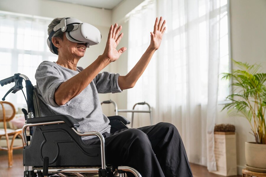 Foto de um homem idoso em uma cadeira de rodas. Ele está usando um óculos de realidade virtual e está com as mãos para a frente como se estivesse se exercitando